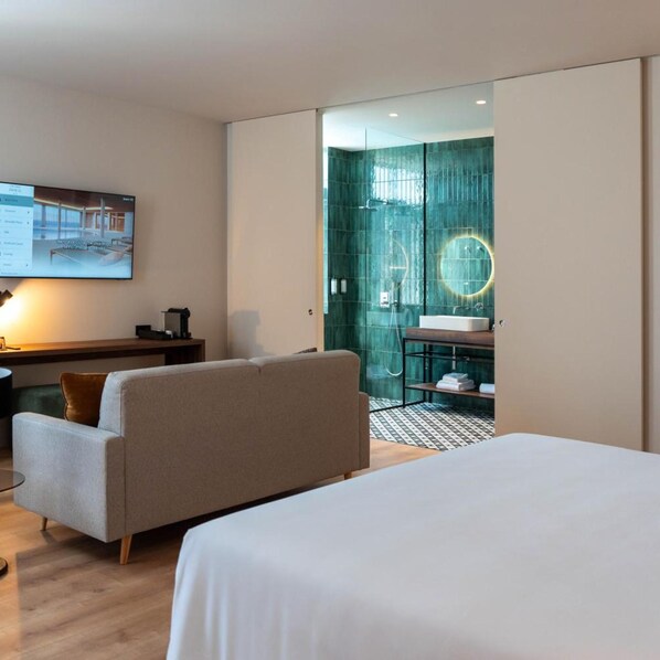 Una habitación de hotel moderna con una cama grande, un sofá, un escritorio con una computadora y un baño con puerta de vidrio.