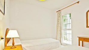 Ein schlichtes Zimmer mit zwei Betten, einem Nachttisch mit Lampe, einem Fenster mit Vorhängen und einer hölzernen Bank.