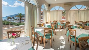 A dining area with tables set for guests, large windows offering a view of the beach and mountains, and a ceiling fan. - Astir Palace (Zakynthos)
