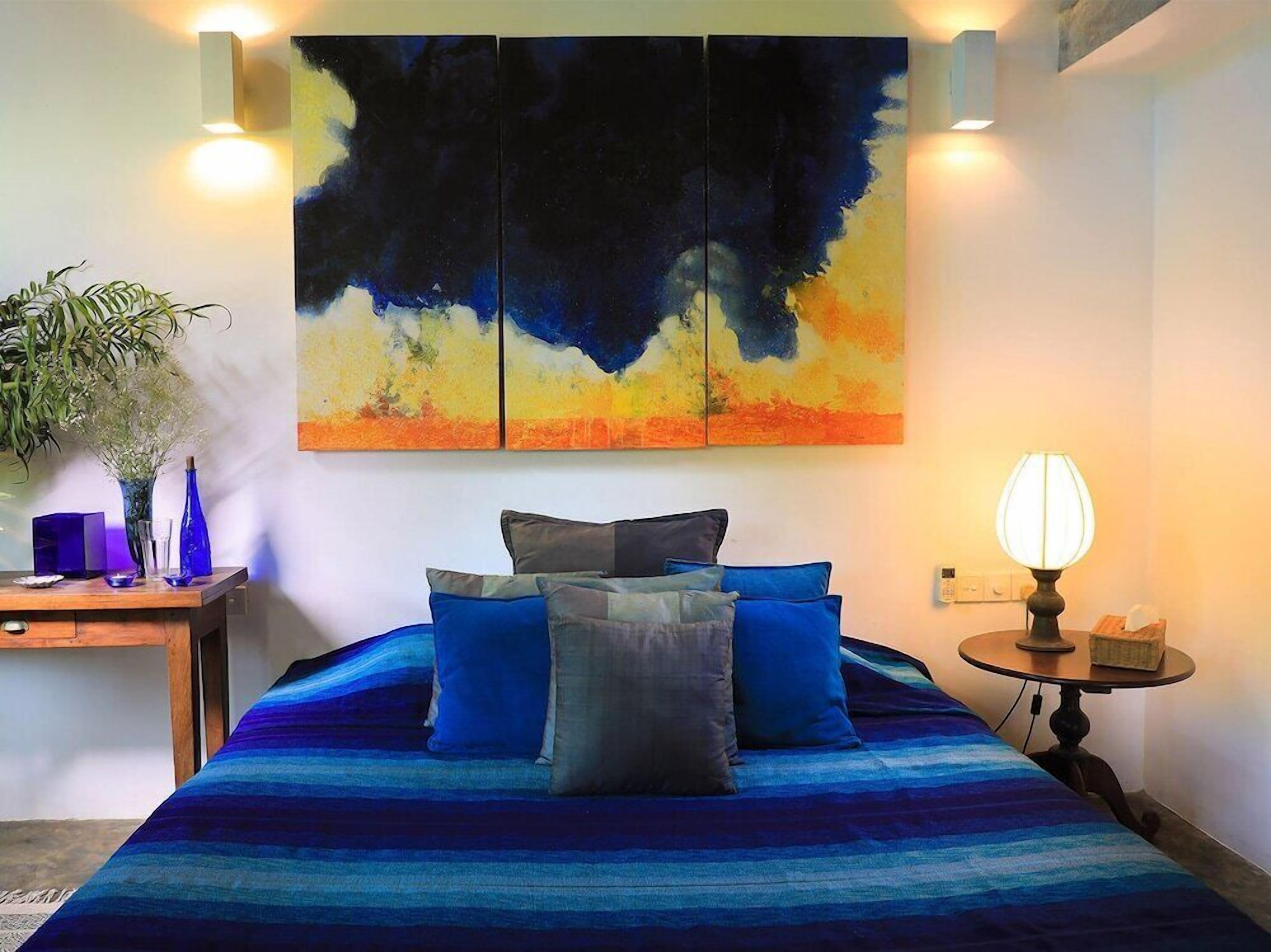 A bedroom with a large bed, a nightstand, a lamp, and a wall-mounted abstract painting.