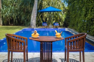 Escena junto a la piscina con dos sillas de madera y una mesa, rodeado de abundante follaje y una sombrilla azul.