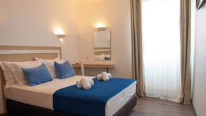 A neatly made bed with a blue blanket, white pillows, and two rolled towels. There is a bedside table with a lamp and a mirror above it. The room has wooden flooring and beige curtains on the window. - Albufeira Beach Hotel (Albufeira)