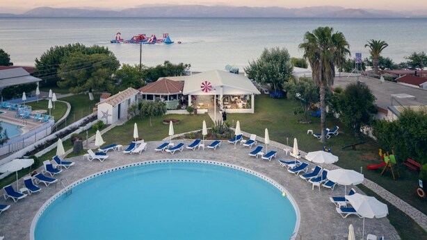 A pool area with lounge chairs and umbrellas, a small building with a red and white striped awning, and a view of the sea. - Albatros Hotel (Corfu)