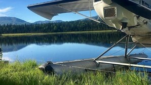 Un petit hydravion est garé sur une zone herbeuse, près d’un lac paisible, avec des montagnes en arrière-plan.