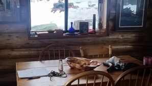 L’intérieur d’un chalet rustique, avec une table à manger en bois, des chaises et une grande fenêtre donnant sur un paysage enneigé.