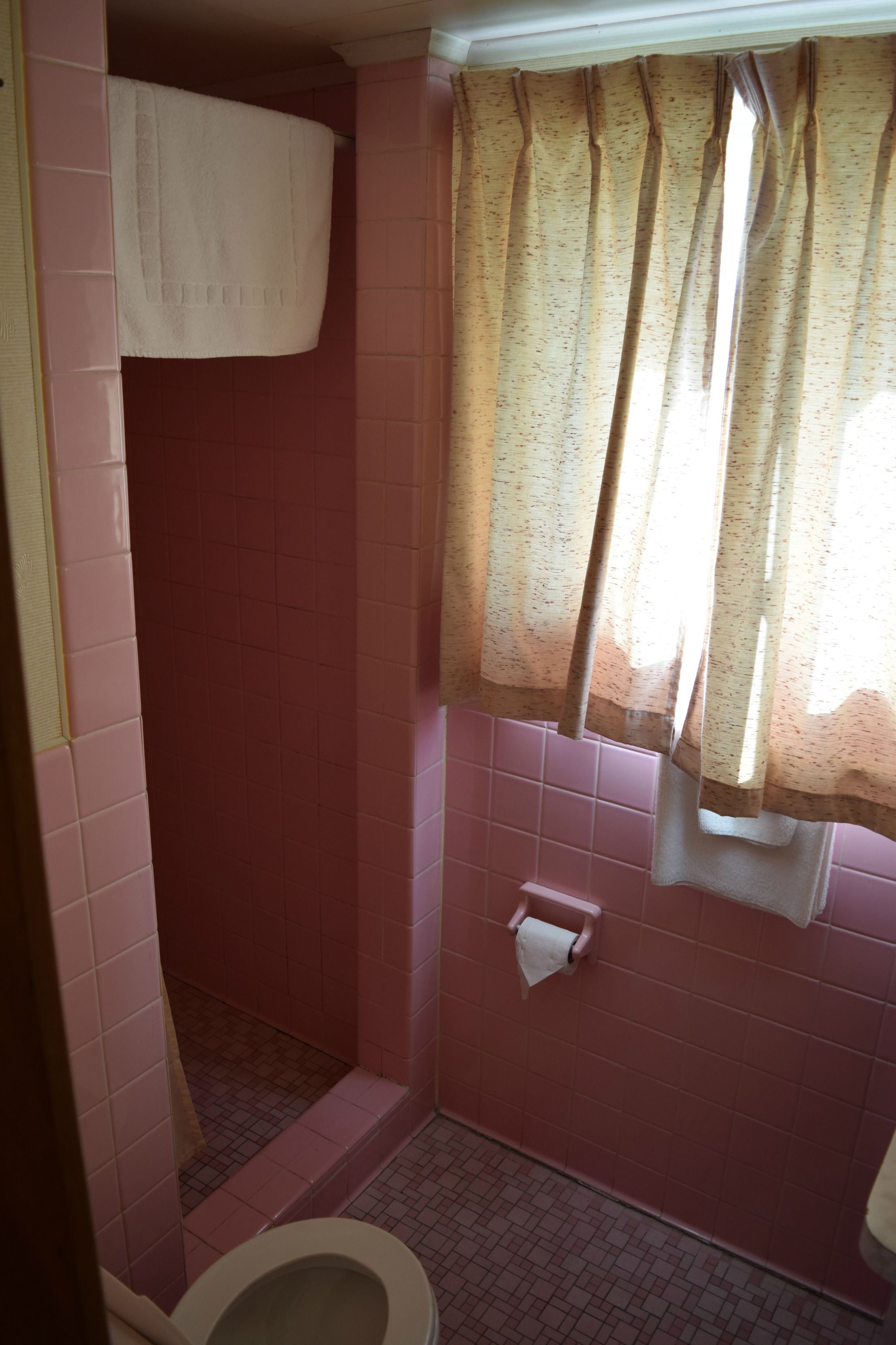 A bathroom with pink tiles, a toilet, a shower, and a window with a curtain.