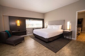 Un hotel moderno con una cama grande, una zona de estar con sofá y sillón, un escritorio y vistas a la ciudad por la ventana.