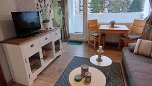 A modern living room with a flat-screen TV, a dining area with a table and chairs, a sofa, and a large window with a view of buildings and trees. - Pure resin romance; Time out for 2 persons; feel good and relax (Altenau)