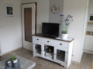 A modern living room with a flat-screen TV on a wooden cabinet, a potted plant, and a small table with two cups.