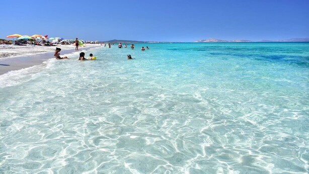 Een strand met helder turquoise water, mensen die zwemmen en zonnebaden, en parasols op de achtergrond.