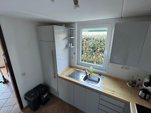 Una cocina compacta con armarios blancos, encimera de madera, ventana con vistas a la vegetación y un cubo de basura.
