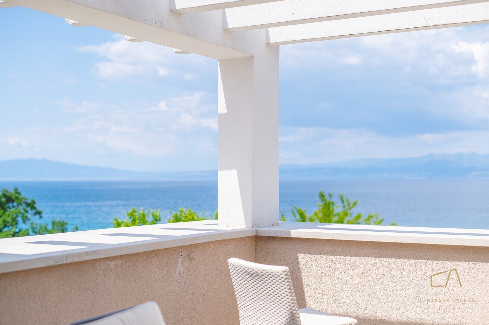 A balcony with a view of the sea and mountains, white wicker furniture, and a textured wall with a logo.