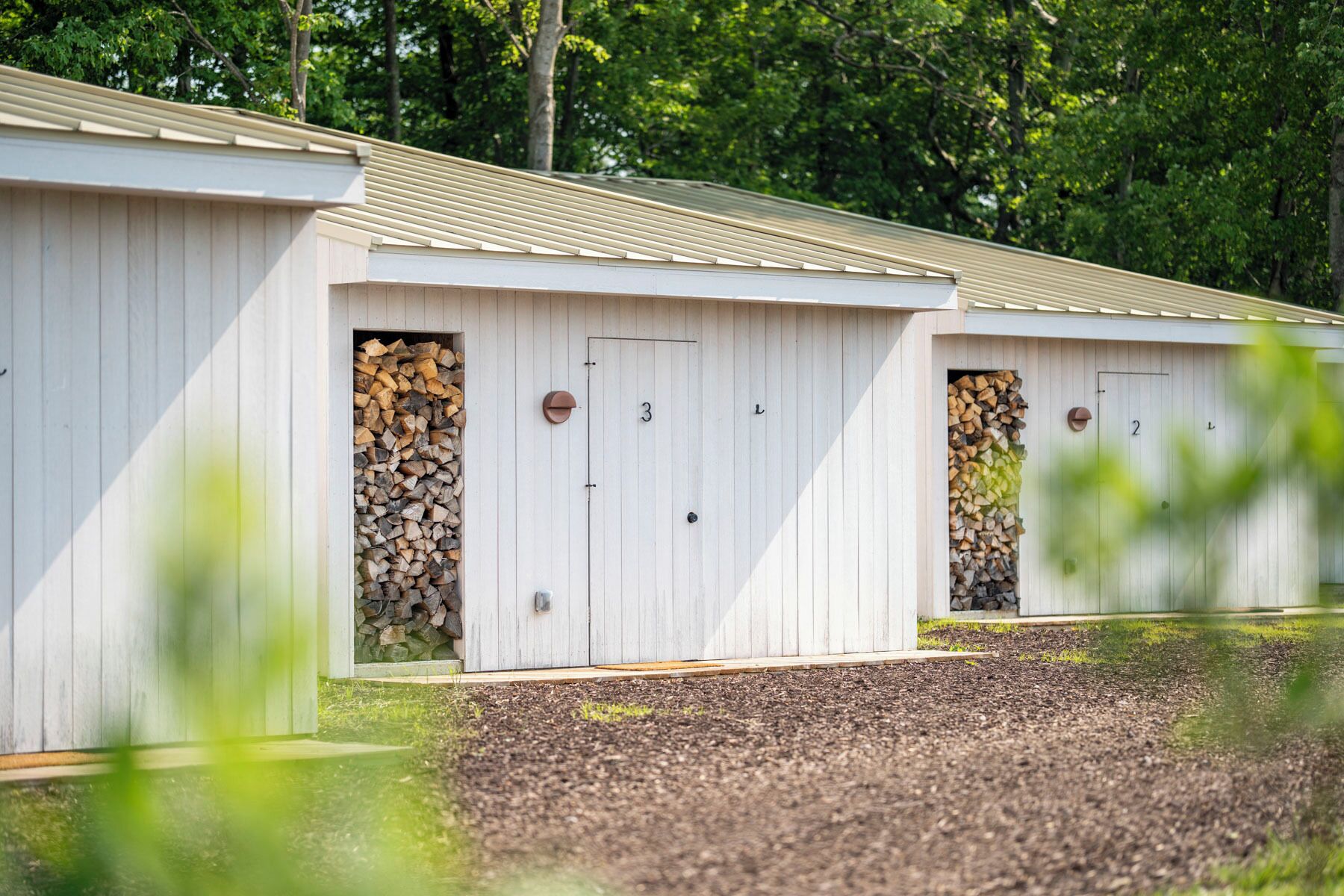 Een rij witte houten huisjes met genummerde deuren, elk met een stapel houtblokken erin.
