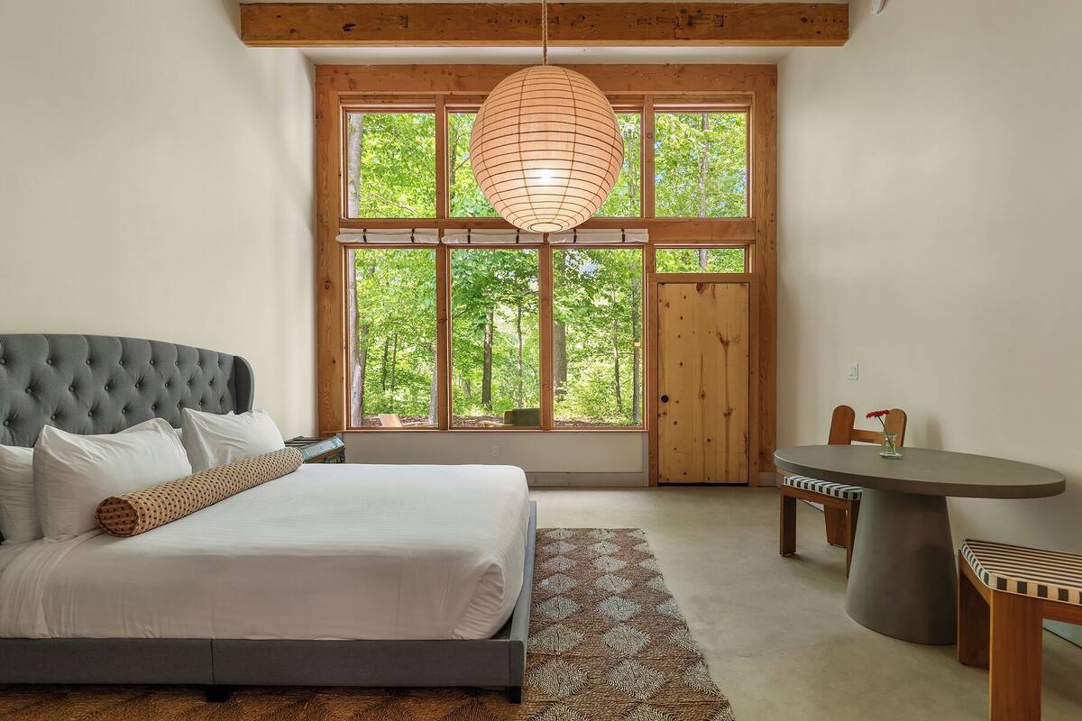 Un dormitorio con una cama grande, techo de madera, ventana con vista a árboles y una pequeña mesa con sillas.