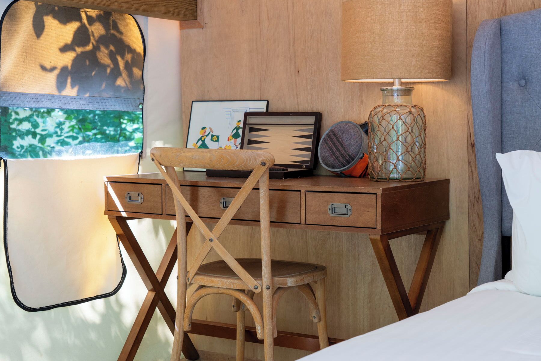 A wooden desk with a laptop, a framed picture, a lamp, and a chair in a room with a window and a bed.