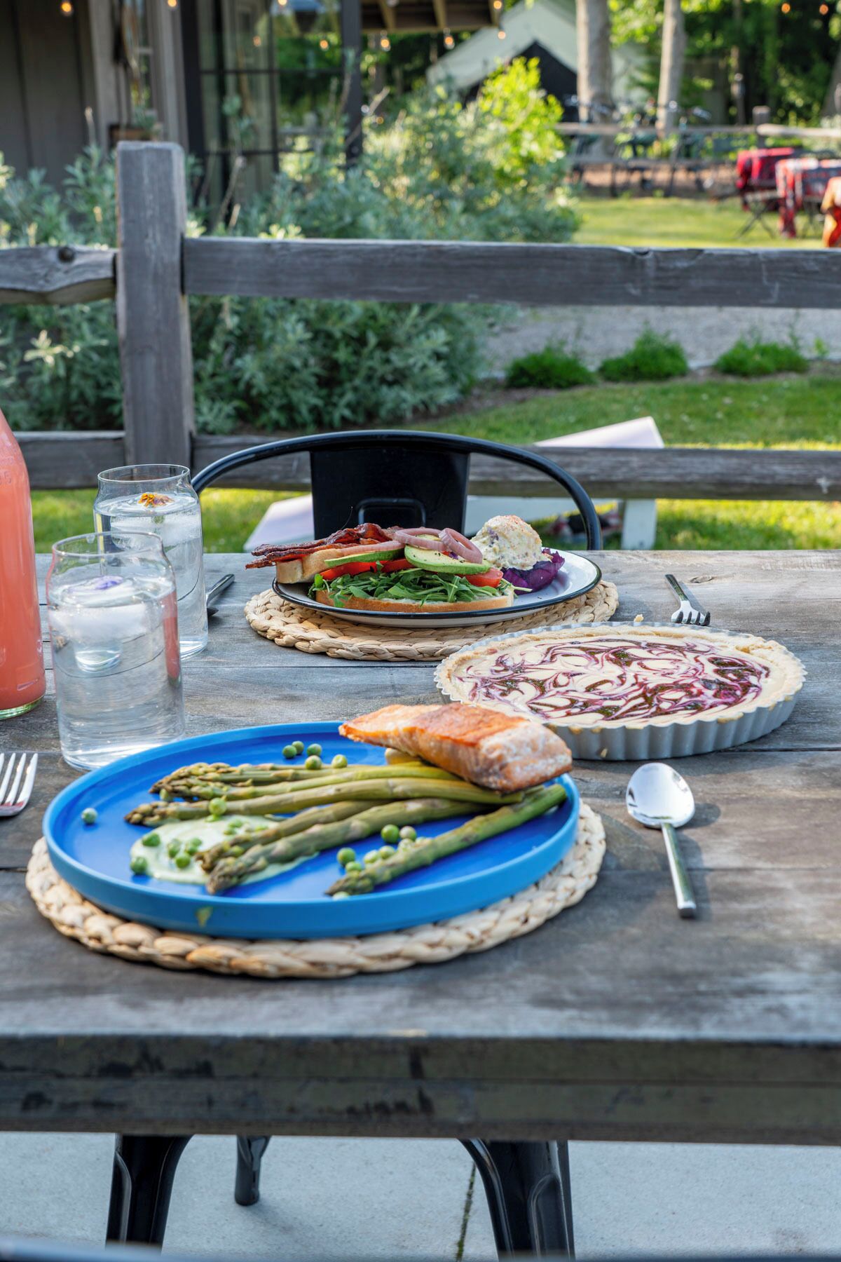 A picnic table with plates of food, including asparagus, salmon, and a pie, set outdoors.