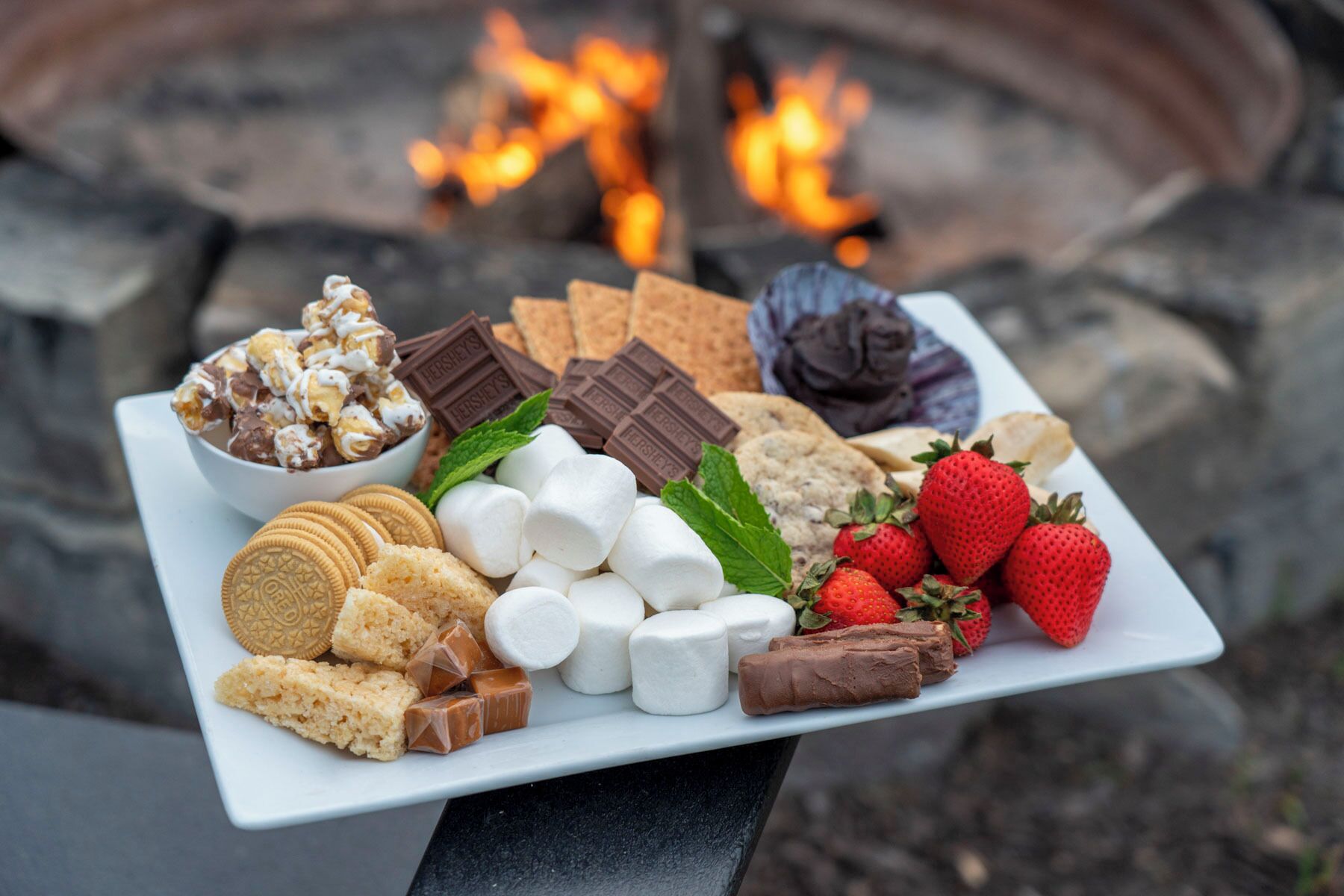 A tray of assorted snacks including marshmallows, strawberries, cookies, and chocolate bars, set against a backdrop of a fire.