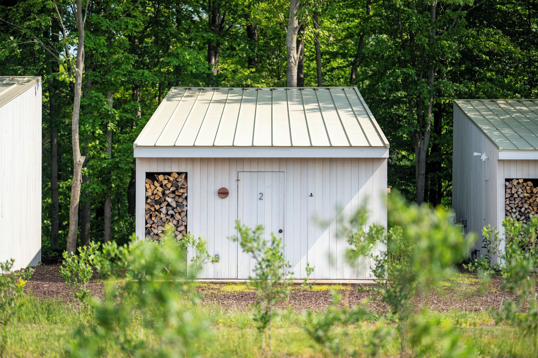 A small, white wooden shed with a metal roof, surrounded by trees and grass.