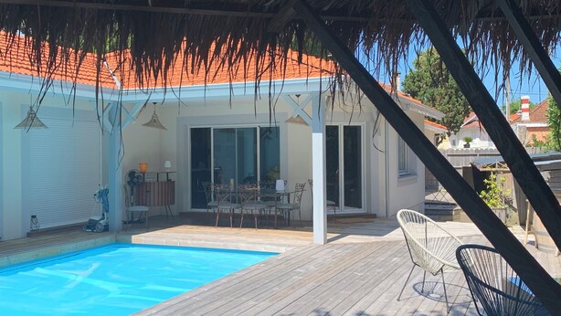 Una terraza de madera con piscina, un porche cubierto y vista a una casa con tejado rojo.