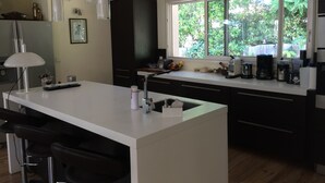 Una cocina moderna con una isla central, taburetes de bar, un amplio ventanal con vistas a zonas verdes y diversos electrodomésticos.