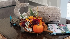 A living room with a glass coffee table holding a decorative arrangement, a woven pouf, and a magazine.