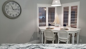 A dining area with a white table, two wicker chairs, a clock, and a window with blinds.