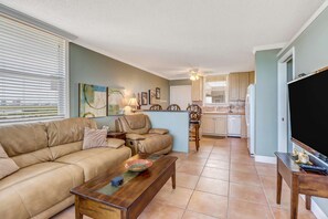 A living room with a beige sofa, a wooden coffee table, and a dining area with a wooden chair and table. A kitchen with white appliances is visible in the background. - DIRECT GULF VIEW | 10TH FLOOR BALCONY |  KING BED | FREE PARKING | STEPS TO BONITA BEACH (Bonita Springs)