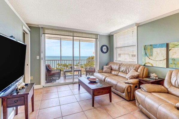 A living room with a large TV, leather sofas, a coffee table, and a view of the ocean through a sliding glass door.