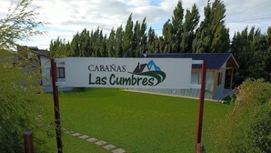 A sign for Cabanas Las Cumbres with a view of a grassy area and a building in the background. - Cabañas Las Cumbres (El Calafate)