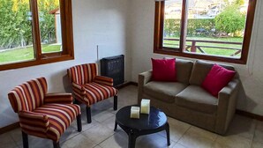 A living room with striped armchairs, a sofa with pink cushions, and a small table with candles. Large windows offer a view of a garden and buildings outside. - Cabañas Las Cumbres (El Calafate)