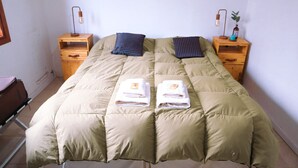 A neatly made bed with a beige duvet, two pillows, and two folded towels on the bed. There are two wooden nightstands with lamps and a small plant on one. - Cabañas Las Cumbres (El Calafate)