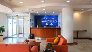 A hotel reception area with a blue sign reading "HOTEL Misión," a reception desk, a plant, and orange seating. - Mision Veracruz (Boca del Río)