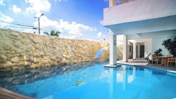 A swimming pool with a stone wall backdrop, a covered seating area, and a clear blue sky. - Mision Veracruz (Boca del Río)