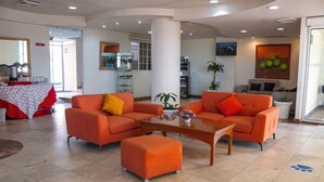 A hotel lobby with orange sofas, a coffee table, and a reception area with a counter and kitchen appliances in the background. - Mision Veracruz (Boca del Río)