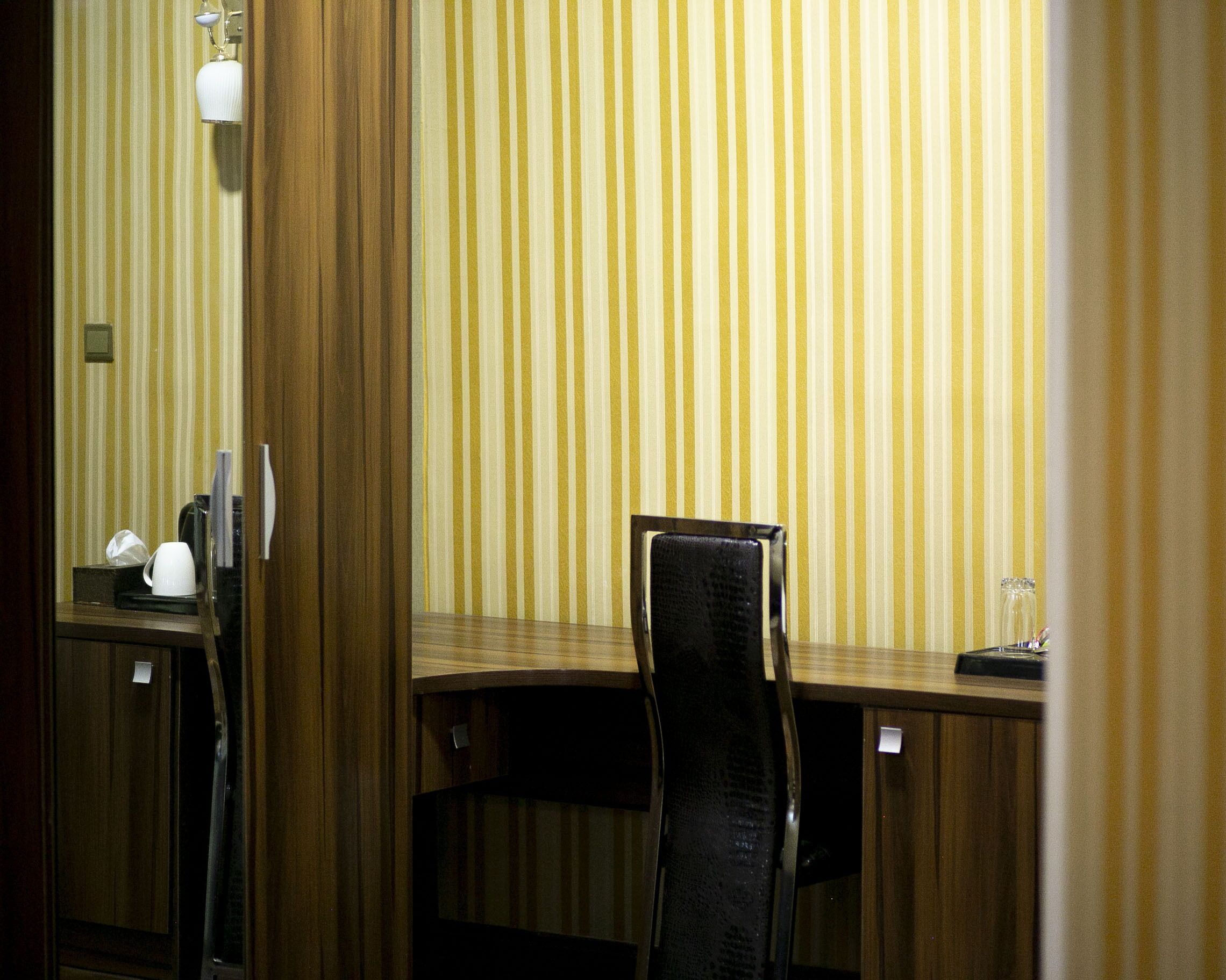 A hotel room with a striped wallpaper, a wooden desk, a black chair, and a bedside table with a lamp and a cup.