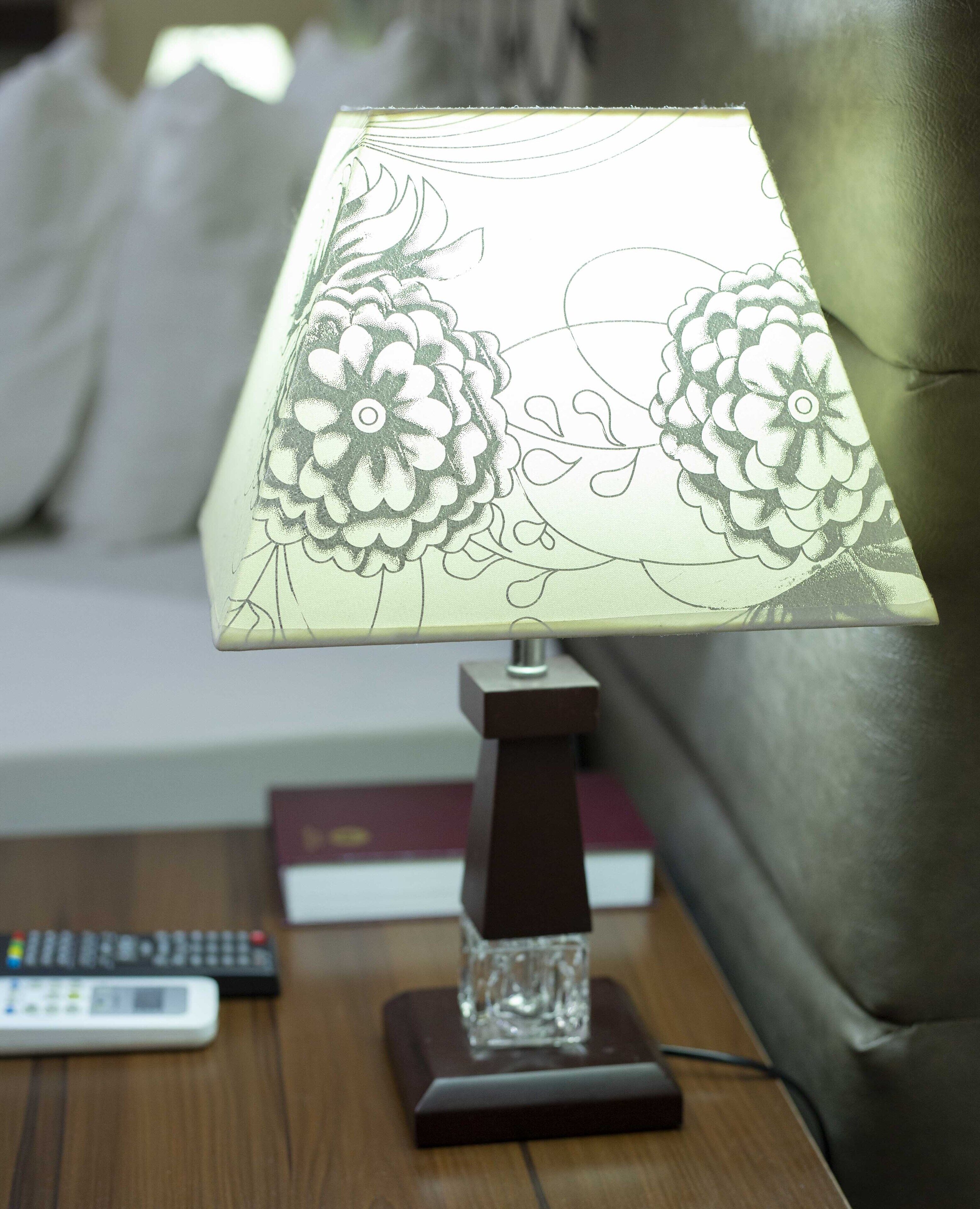 A table lamp with a floral design on its lampshade, placed on a wooden nightstand next to a remote control and books.