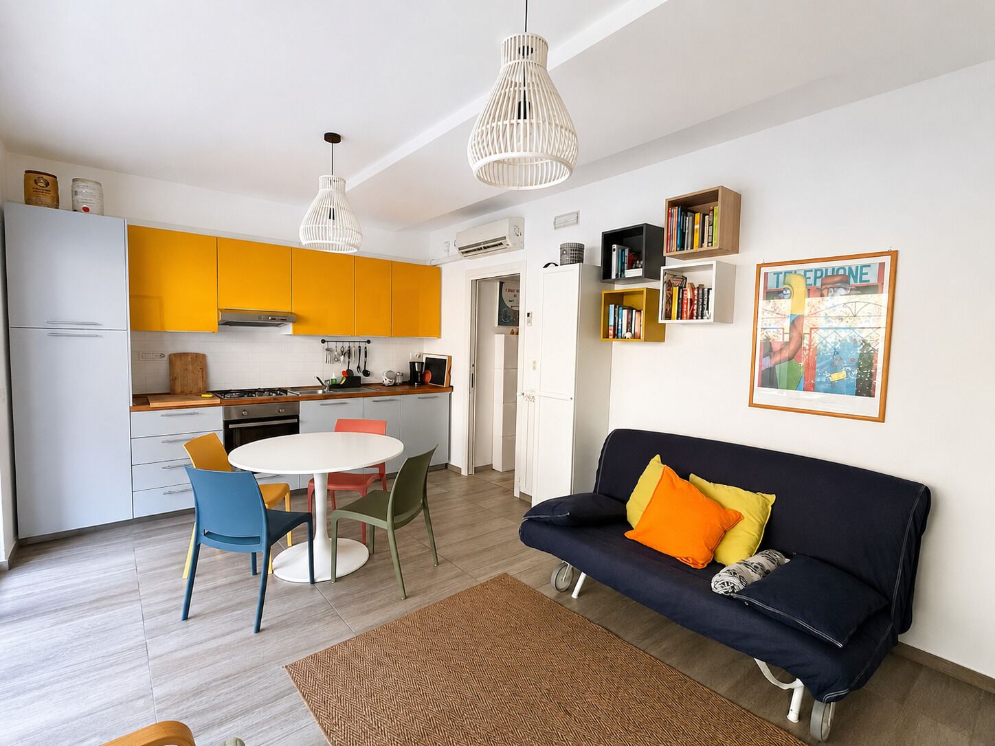 A modern kitchen with yellow cabinets, a dining table with chairs, a black sofa with orange cushions, and a bookshelf.