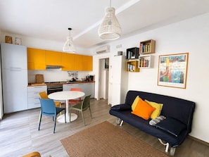 A modern kitchen with yellow cabinets, a dining table with chairs, a black sofa with orange cushions, and a bookshelf.