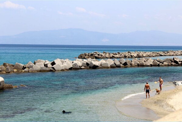 A beach with rocky breakwaters, clear blue water, and distant mountains.