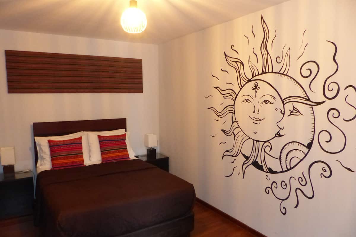 Un dormitorio con cama, mesitas de noche, una lámpara de pared y un mural en la pared.