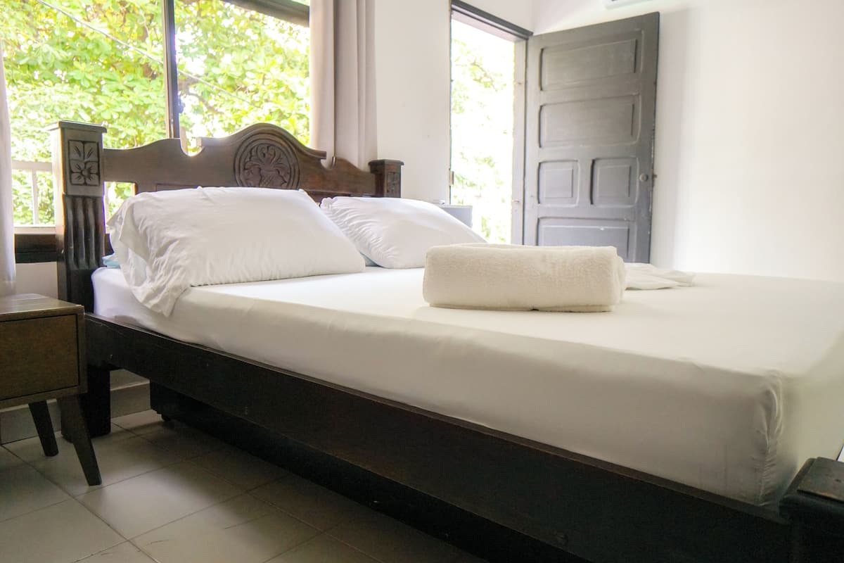 A neatly made bed with white linens and a folded towel, a wooden nightstand with a lamp, and a window with a view of greenery.