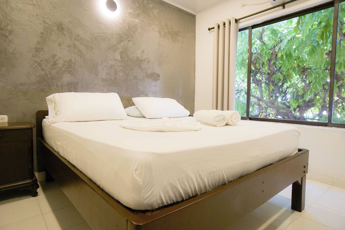A neatly made bed with white linens and pillows, a wooden headboard, and a window with curtains.