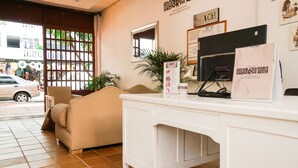 A reception area with a beige sofa, a white counter, and a computer monitor. There is a window with a view of the street outside. - Hostal Paraiso Tayrona (Santa Marta)