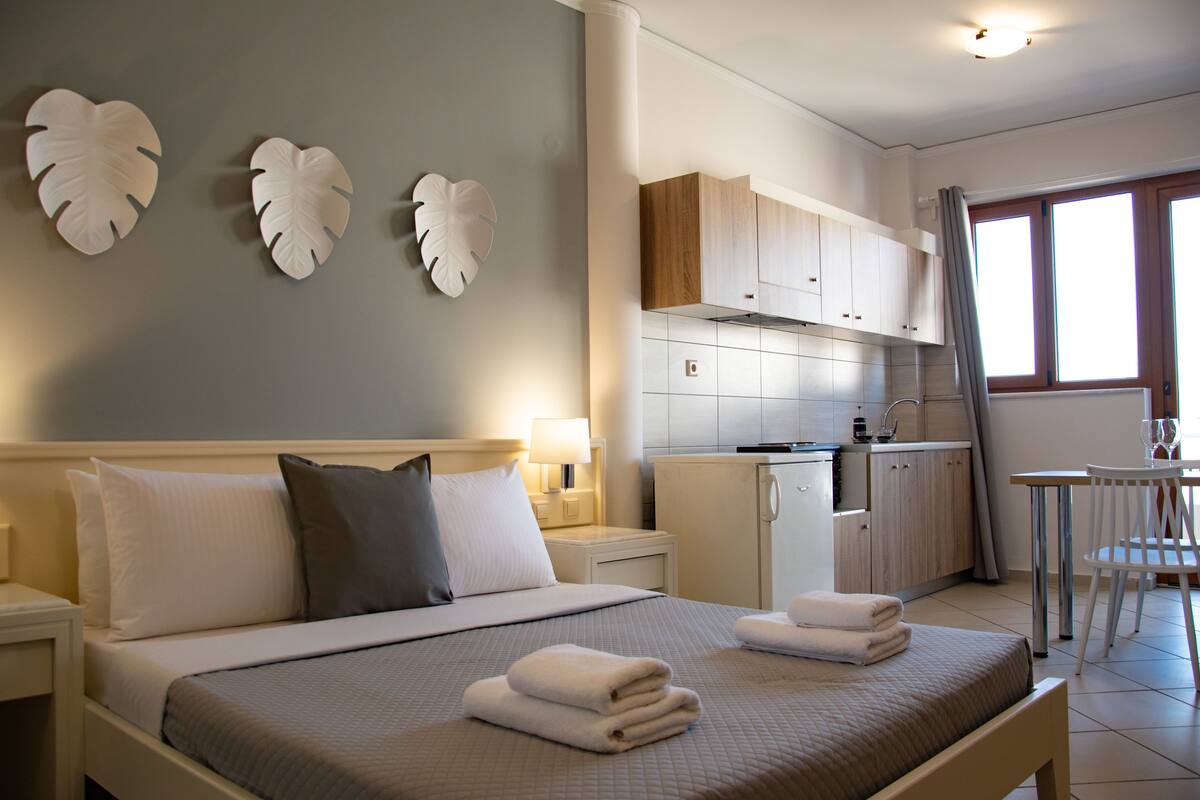 Una habitación de hotel con cama, mesitas de noche, escritorio, silla, kitchenette y ventana.