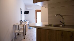 A modern kitchen with wooden cabinets, a stainless steel sink, and a white countertop. There is a dining area with a white chair and a small table. - Horizon Apartments Platanias (Platanias)