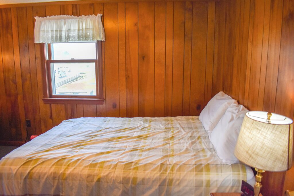 Ein Schlafzimmer mit Holzwandverkleidung, einem Fenster mit weißem Vorhang und einem Bett mit weißer und gelber Bettdecke sowie einer Nachttischlampe.