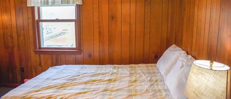 Ein Schlafzimmer mit Holzwandverkleidung, einem Fenster mit weißem Vorhang und einem Bett mit weißer und gelber Bettdecke sowie einer Nachttischlampe.