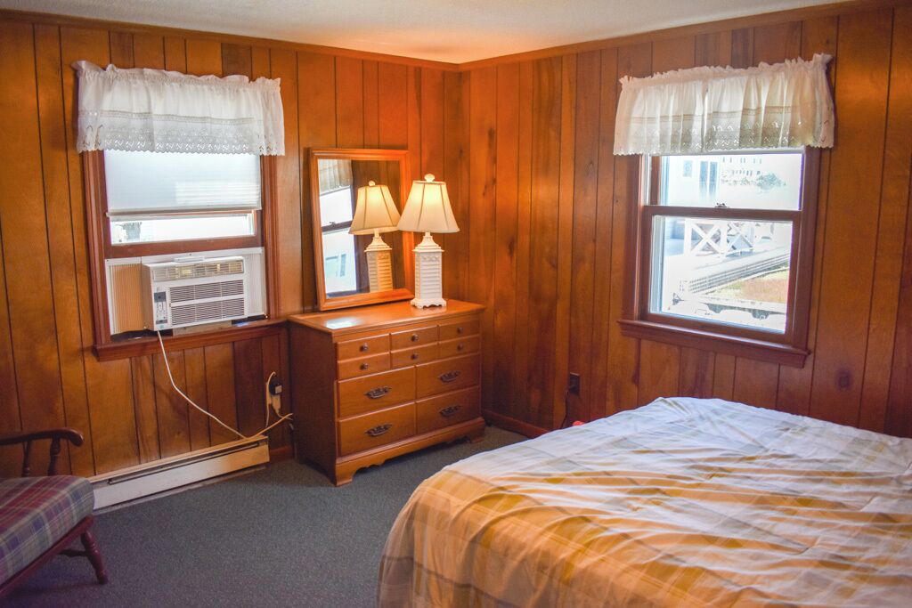 A bedroom with wooden paneling, a bed, a dresser with lamps, and a window with a view of a train track.