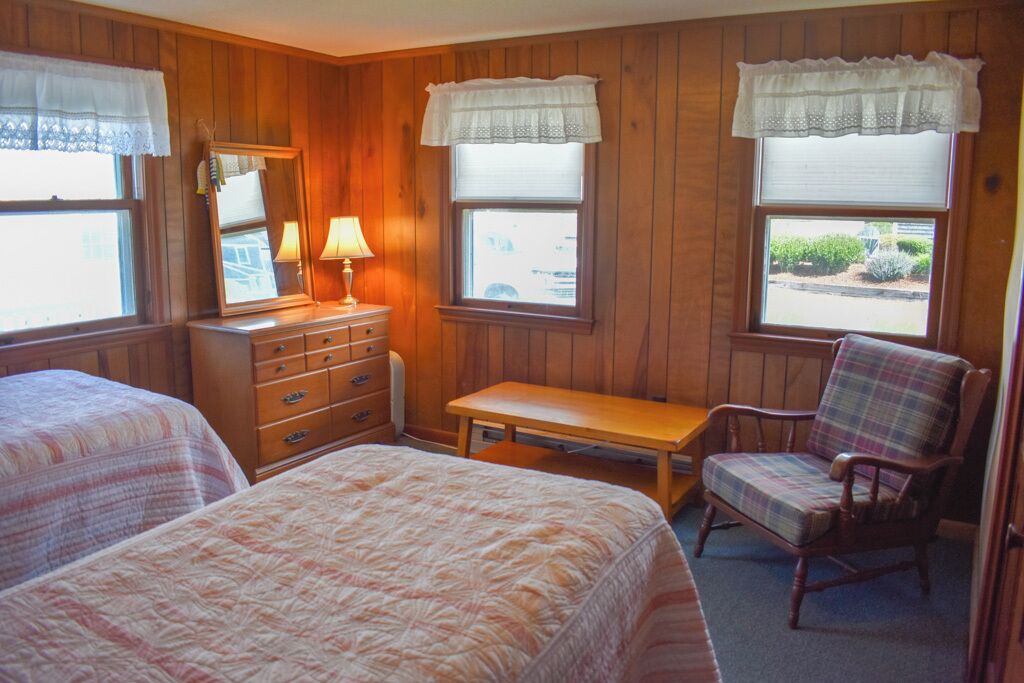 A room with two beds, a dresser, a chair, and windows with curtains.
