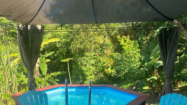 A poolside area with a wooden deck, two blue chairs, a table with food and drinks, and a pool with a blue interior. - TropicalKaz Guadeloupe - Villa Sunset (Sainte Rose)